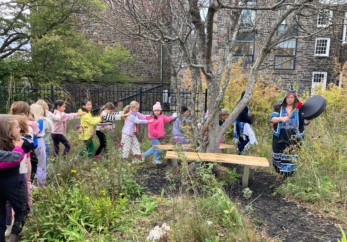 A line of childrean walking through a garden following a Mi'kmaw woman in traditional regalia playing a hand drum.