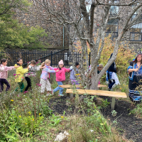 A line of childrean walking through a garden following a Mi'kmaw woman in traditional regalia playing a hand drum.